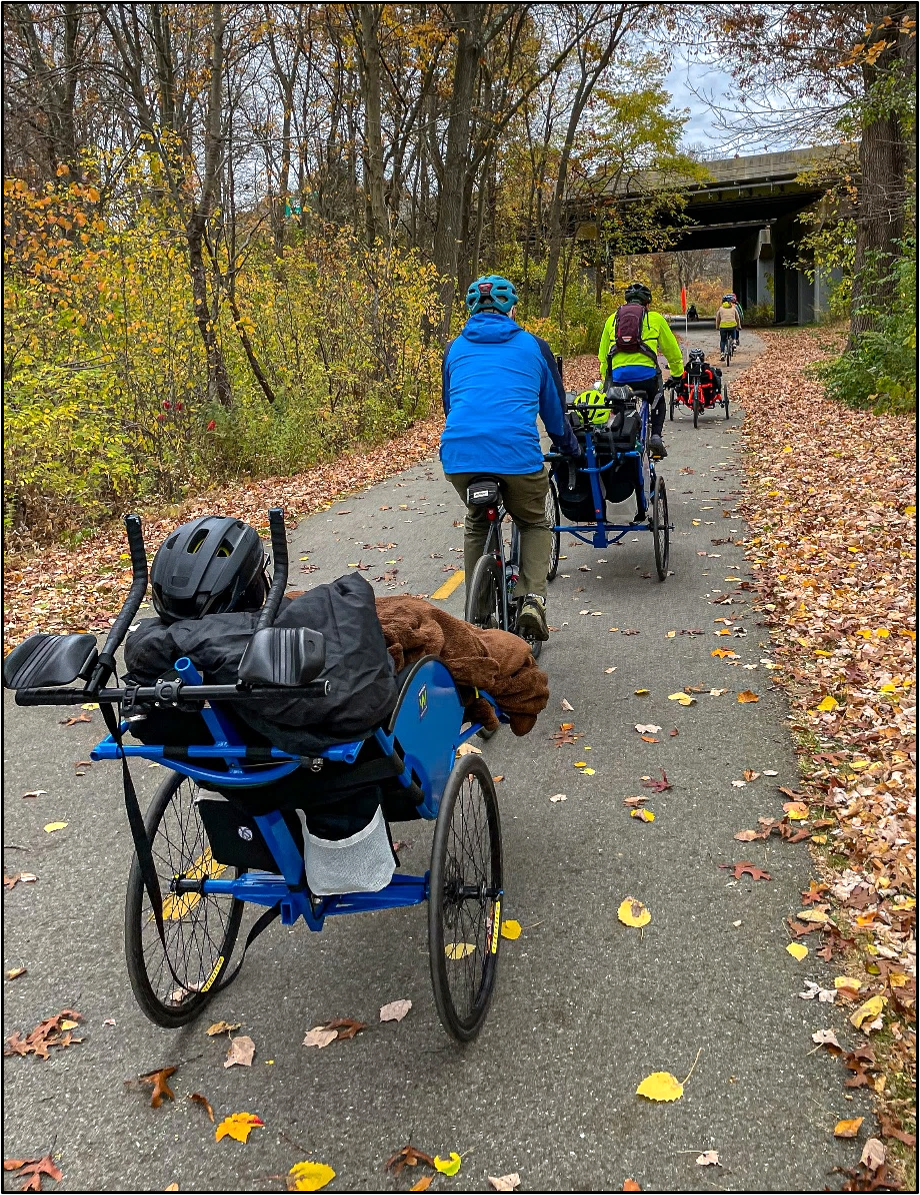 A group of people ride a variety of tandem adaptive cycles  along a paved bike path in autumn. 