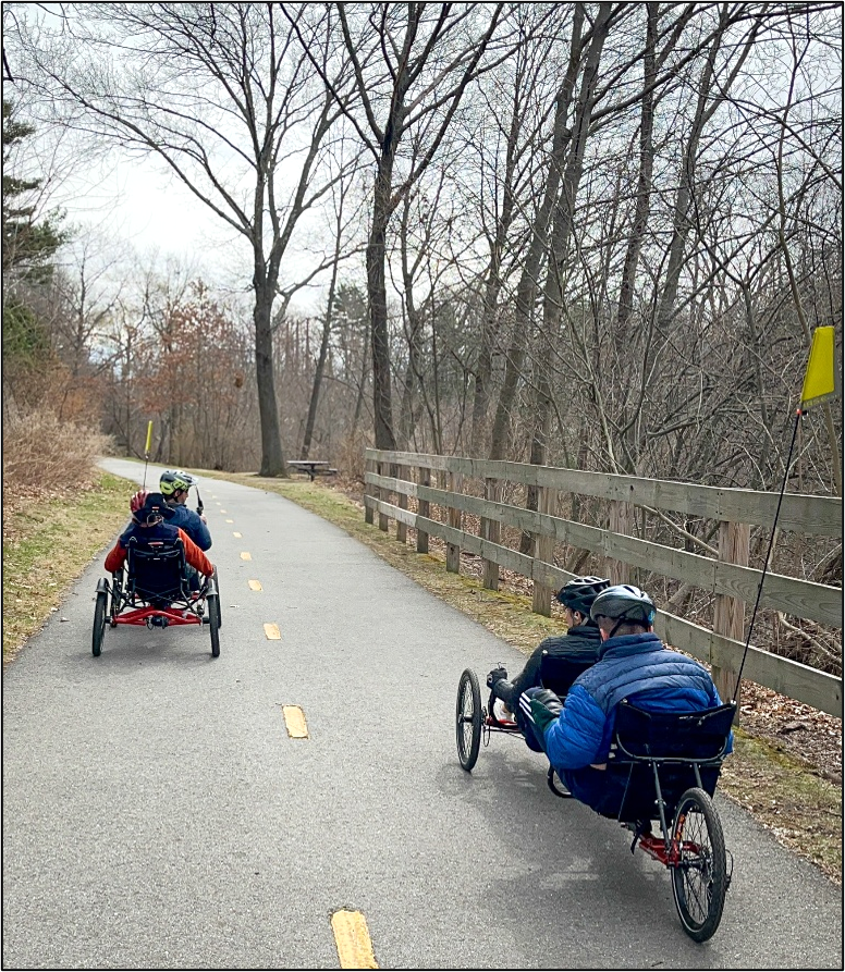 People are peddling 2 recumbent tandem cycles along a paved bike path in autumn.