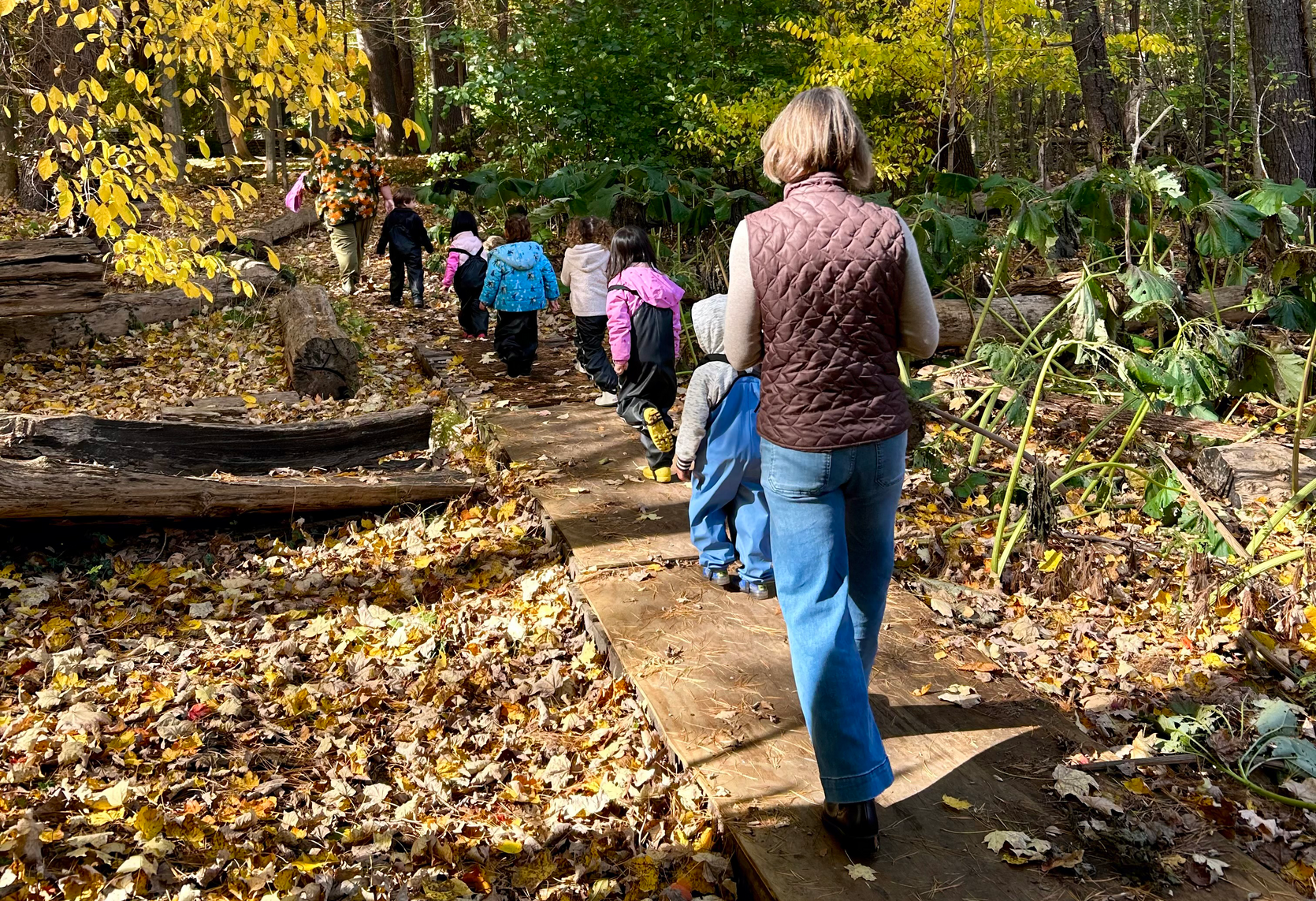 An educator and small children walk away from the camera in single file outside in a wooded area.
