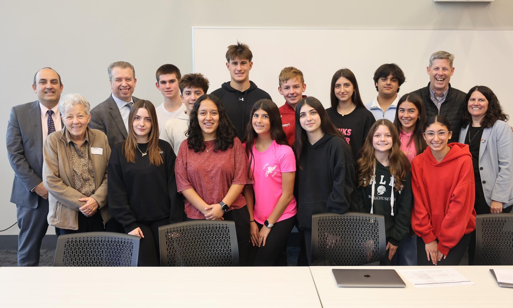 A group of educators and high school students smile and pose for a group photograph in a white conference room. A table and chairs are visible in the foreground. 
