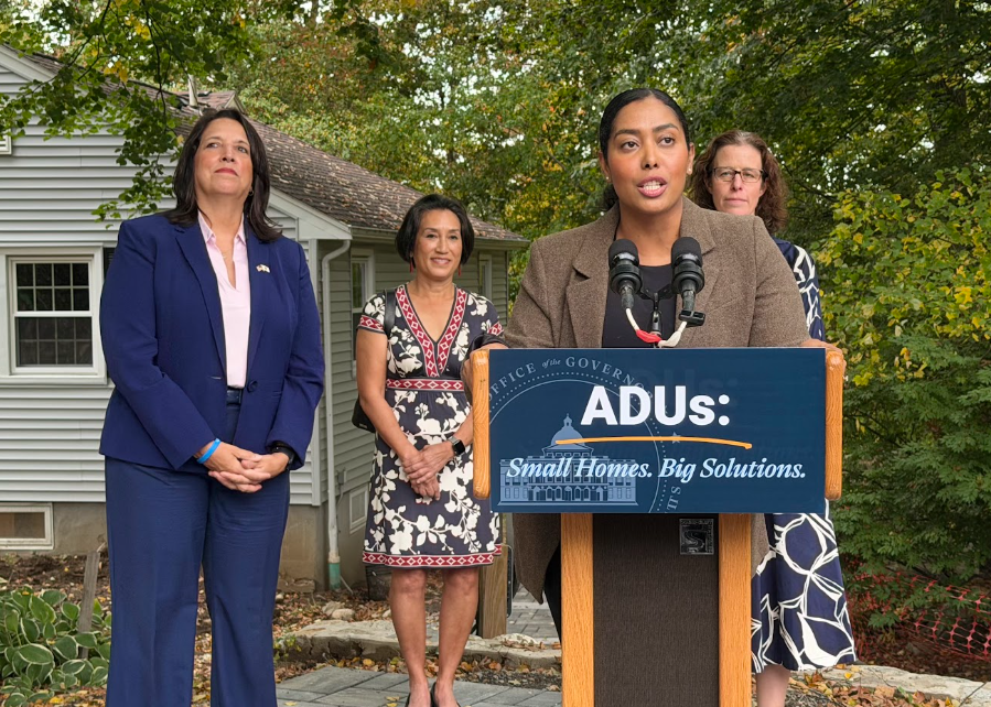 Secretary Juana Matias stands at a podium in front of a home with Lieutenant Governor Kim Driscoll beside her.