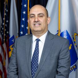 Secretary Zrike stands in front of a U.S. and Massachusetts state flag wearing a grey suit and blue tie. 