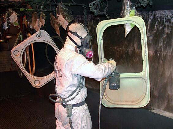 A person in a white full-body suit and respirator paints an automotive part.