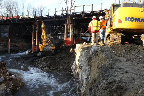 Construction equipment and workers on a concrete wall near rushing water.
