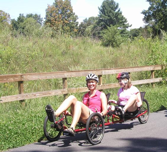 Two riders on a Terra Trike Rover Tandem peddling on a rail-trail near a grassy field.