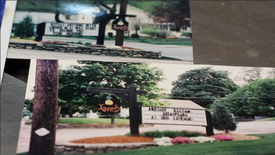 An old photograph sits on a table in Tom's woodshop. In the photo is a sign outside of Xavier's Restaurant. Writing on the sign says "Another section disappears at our expense". Another photo of the same sign sits on the table off-center. The writing is blurry, but the name Tom Hopkins can be seen.