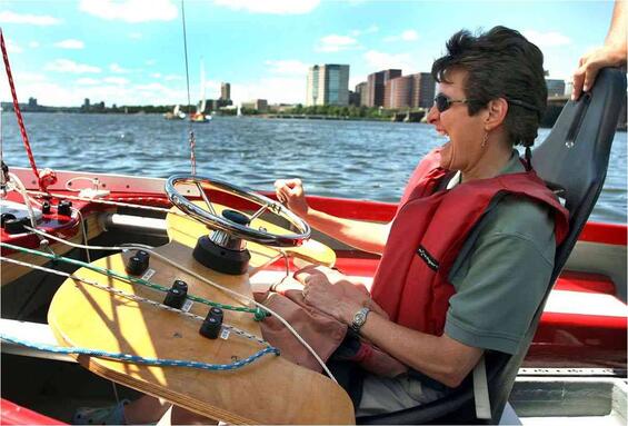 A woman is sailing in a seat with high back support. In front of her is a flat console with a wheel and cleats holding colored lines.