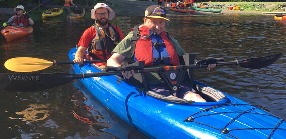 A paddler in the front of a tandem kayak has their paddle clipped into a Y-shaped bracket that stands up in the boat in front of their seat. The bracket holds the paddle at the right height for paddling.
