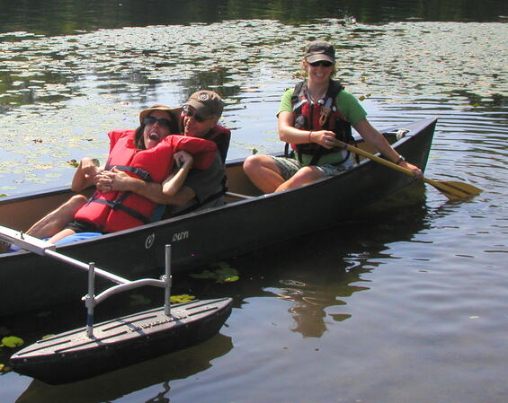 A paddler sits in the lap of another paddler on the hull of a canoe with outriggers. Another paddler holding a paddle sits on the stern seat of the canoe.
