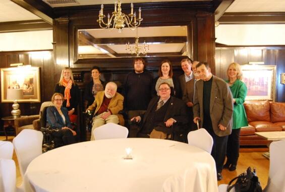 Tom poses with 10 colleagues in front of a fireplace. A chandelier is above them and a table draped in a white tablecloth is in front of them.