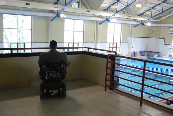 Tom sits on a balcony overlooking an indoor pool. His back is to the camera.