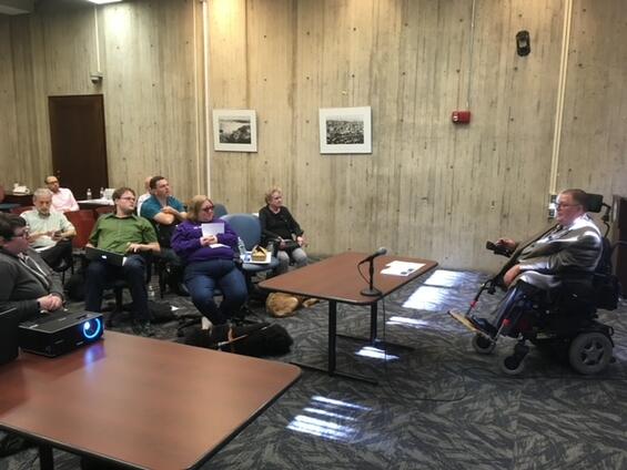 Tom sits at a table with a microphone presenting to a group of people. They are indoors and a projector is visible on the table next to Tom.