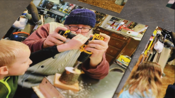 Old photograph of Tom sitting at a work table in his wood shop. A small, short section of a narrow tree trunk sits on the table on top of a wooden base in front of him. Tom is using both hands to hold a drill drilling a hole into the middle of the trunk. His grandson, a small child, stands next to him and also has his hand on the drill.