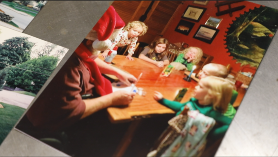 Tom, his back to the camera, sits at a wooden dining table with a Santa hat on. His five grandchildren, all toddlers and young children, sit around the table with him. They are all wearing pajamas and looking intently at a paper Tom is holding in his hands.