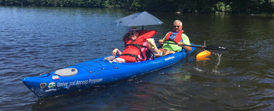 Two paddlers in a tandem kayak with outriggers. The paddler in back is holding a paddle. The paddler in front has an umbrella.