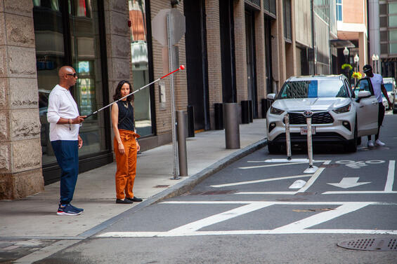 Man with his white cane raised as he stands on a sidewalk in front of a crosswalk while his instructor looks on