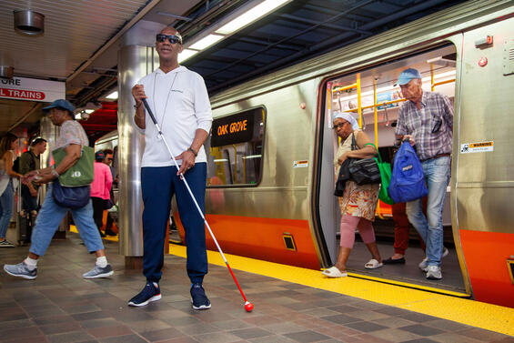 Man standing on a subway platform with his white cane and an MBTA Orange Line train behind him