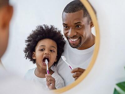 A little kid brushing their teeth and their dad holding a toothbrush looking proud and happy