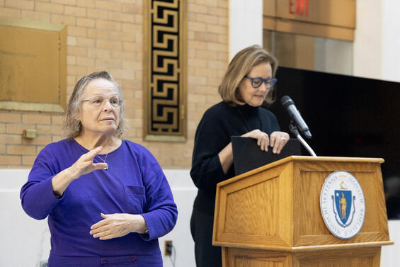 A woman in a black turtleneck shirt speaks at the podium. An ASL interpreter is signing to her left.