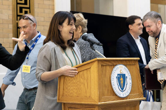 A smiling woman wearing a gray blazer speaking at the podium during the ceremony.