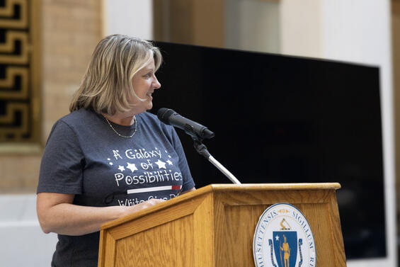 A blonde woman speaking from the podium wearing a blue t-shirt with the text: A galaxy of possibilities. White Cane Day.