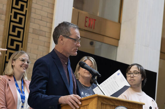 A man in a suit reading from a folder while behind the podium while three people stand behind him watching.