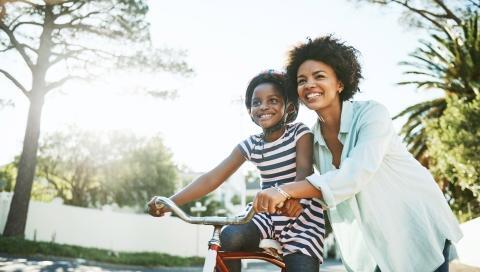A mother is assisting her daughter on a bike. They are both smiling and enjoying the outdoor time together.