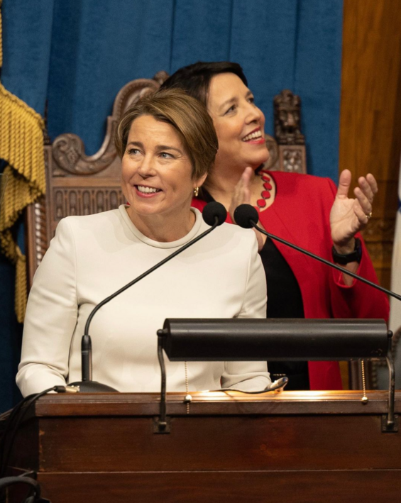 Governor Healey smiling as she delivers her inaugural address. Lt. Governor Driscoll in the background of the picture clapping. 