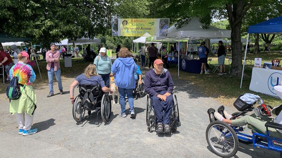 People in mobility devices visit tents set up along a stone dust path in a park.