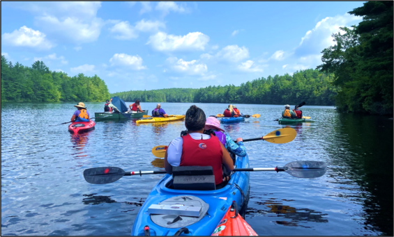 A group of paddlers in brightly colored kayaks on a lake surrounded by trees.