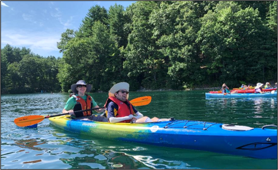 Two people are smiling while paddling in a blue and yellow tandem kayak on a pond.