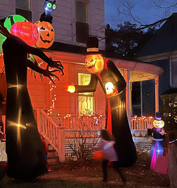 Trick-or-treaters approaching a home decorated with tall pumpkin-headed figures and orange lights for Halloween.