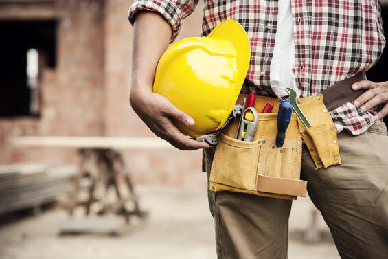 Construction worker holding a yellow hardhat and wearing a tool belt.