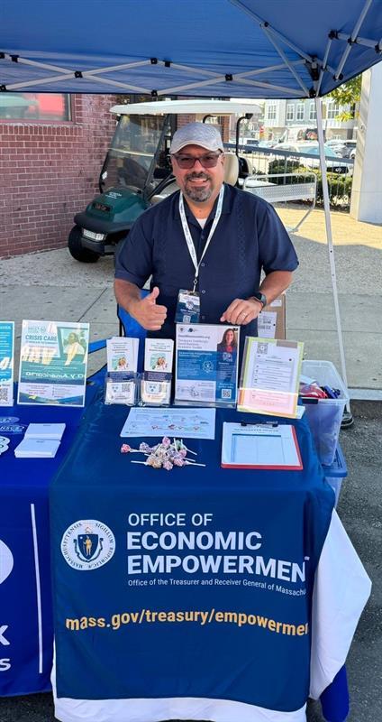 An image of Community Ambassador Jaime Jimenez, smiling and giving a thumbs up, while promoting the Office of Economic Empowerment at a community event. Jaime is standing outdoors behind a table with promotional materials and a blue tablecloth with the logo for the Office of Economic Empowerment.