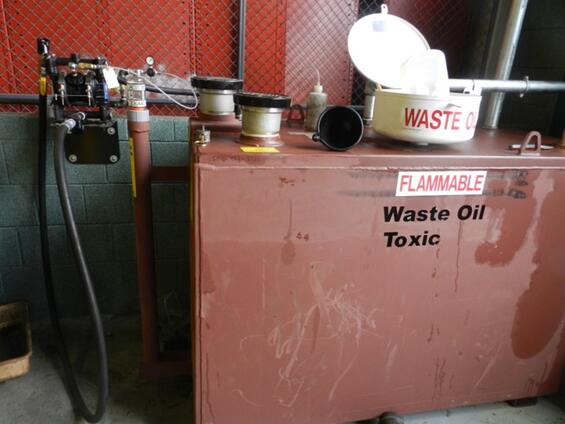 A red waste oil storage tank against a cement wall. Signs on the tank say "WASTE OIL," "FLAMMABLE," and "Waste Oil Toxic."