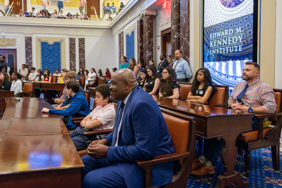 Massachusetts Education Secretary Patrick Tutwiler sits with students and educators in a full-scale replica of the U.S. Senate Chamber at the Edward M. Kennedy Institute in Boston during the Massachusetts Civics Project Showcase. The room is filled with young people and community members, reflecting civic engagement and learning. A large screen in the background displays the Edward M. Kennedy Institute’s name and ornate dome imagery.