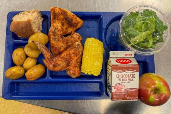 A school lunch tray from Wilmington Public Schools featuring rotisserie chicken wings and breast, roasted baby potatoes, a corn on the cob, a whole wheat dinner roll, a small salad in a clear plastic container, an apple, and a carton of Hood chocolate fat free milk. The tray is blue and divided into compartments.