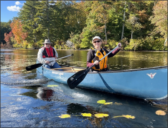 Two people are smiling as they paddle in a canoe on a lake lined with autumn trees. 