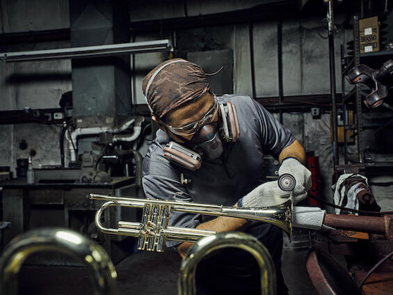 A worker in a respirator, gloves, and goggles works on a trumpet