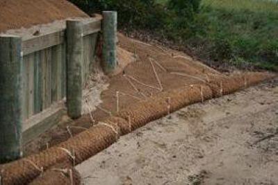 A vertical wooden bulkhead on a sandy bank with fiber blankets around it and coir rolls in front of it