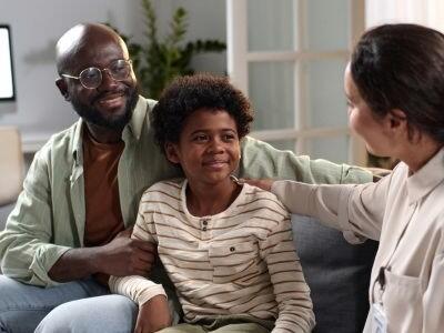 A child sitting with their parent on a couch talking to a Community Health Worker who is holding a clipboard.