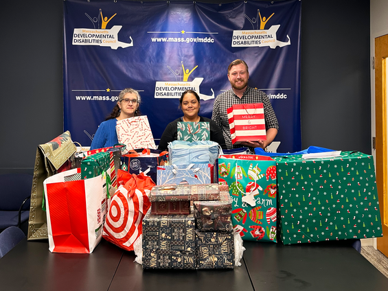 Three people holding holiday gift bags standing behind a pile of gifts.