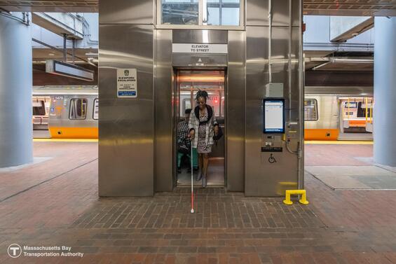 A person with a white cane and a wheelchair user exit the elevator onto the train platform