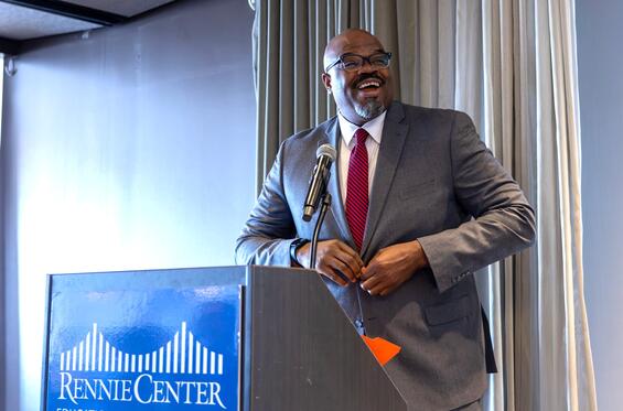 Secretary Tutwiler is wearing a suit with a red tie, standing at a podium with a sign that says Rennie Center