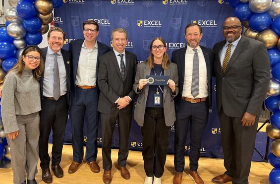 Eight adults stand shoulder to shoulder smiling in front of an Excel Academy Charter Schools step-and-repeat backdrop with blue and gold balloons. At center, a woman wearing glasses holds an award envelope while colleagues in business attire pose alongside her in a school gym.