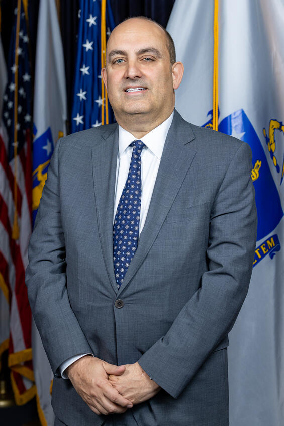 Secretary Zrike in front of the American flag and the Massachusetts state flag.