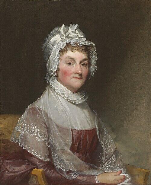 Abigail Adams has a neutral expression with embroidered shawl over her shoulders and a frilly white hat upon her head with a bow. 