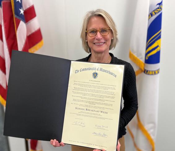 Acting Education Secretary Amy Kershaw stands in front of U.S. and Massachusetts flags, smiling as she holds an open proclamation folder from The Commonwealth of Massachusetts declaring “School Breakfast Week.”