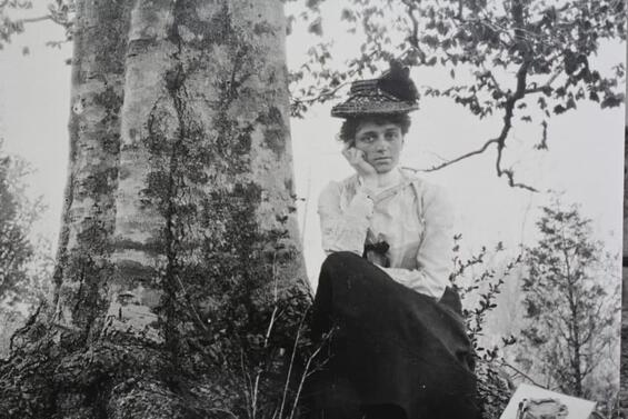 Blanche Ames Ames sitting outdoors with her chin resting on her hand, next to a large tree trunk wearing a long-sleeved blouse with a skirt and a hat.