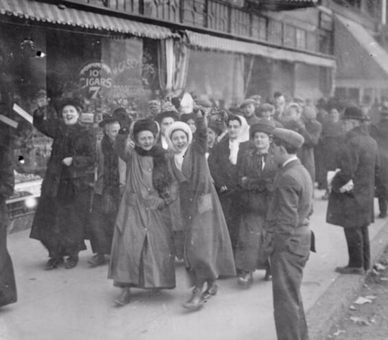 Group of mostly women strikers with one arm raised, wearing winter coats and hats. walking down a city street as mostly women as several med look on.